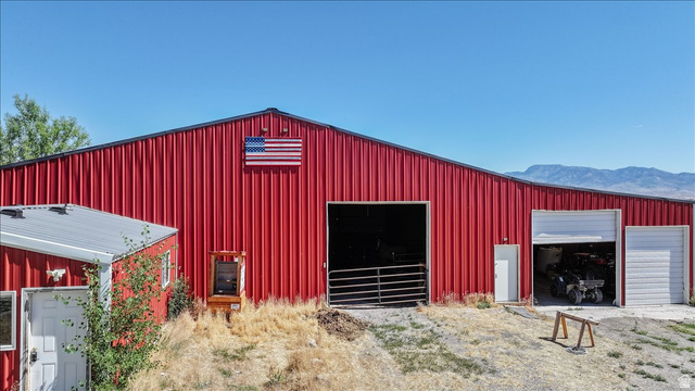 1634 West Arimo Road Arimo, ID 83214 - Photo 60 of 89 Red metal barn with a high-pitch roof, featuring multiple large openings for equipment access, a pedestrian door, and an attached smaller structure with a white overhead door