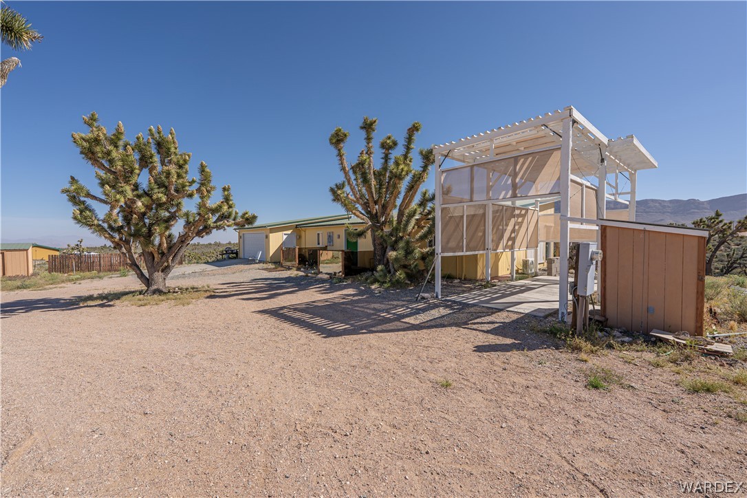 26512 North Yucca Road Meadview, AZ 86444 - Photo 17 of 38 The property features a spacious gravel yard with mature Joshua trees, a yellow-sided building with a garage door, and a white pergola structure with an attached shed