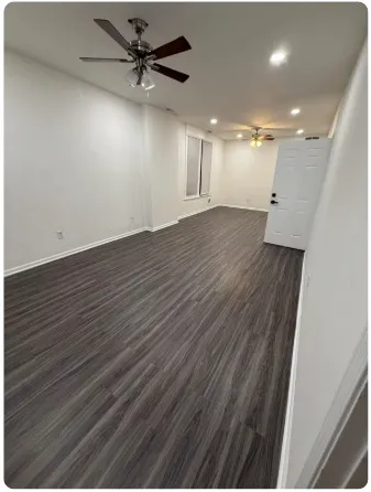 an empty room with wooden floor ceiling fan and kitchen view