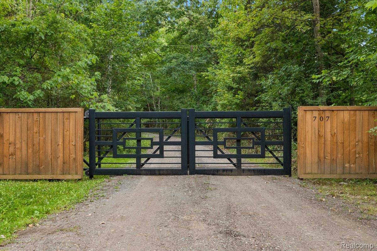 707 West Sutton Road Metamora, MI 48455 - Photo 16 of 84 Wrought iron gate with geometric detailing, flanked by natural wood fencing, opening to a gravel driveway surrounded by mature trees