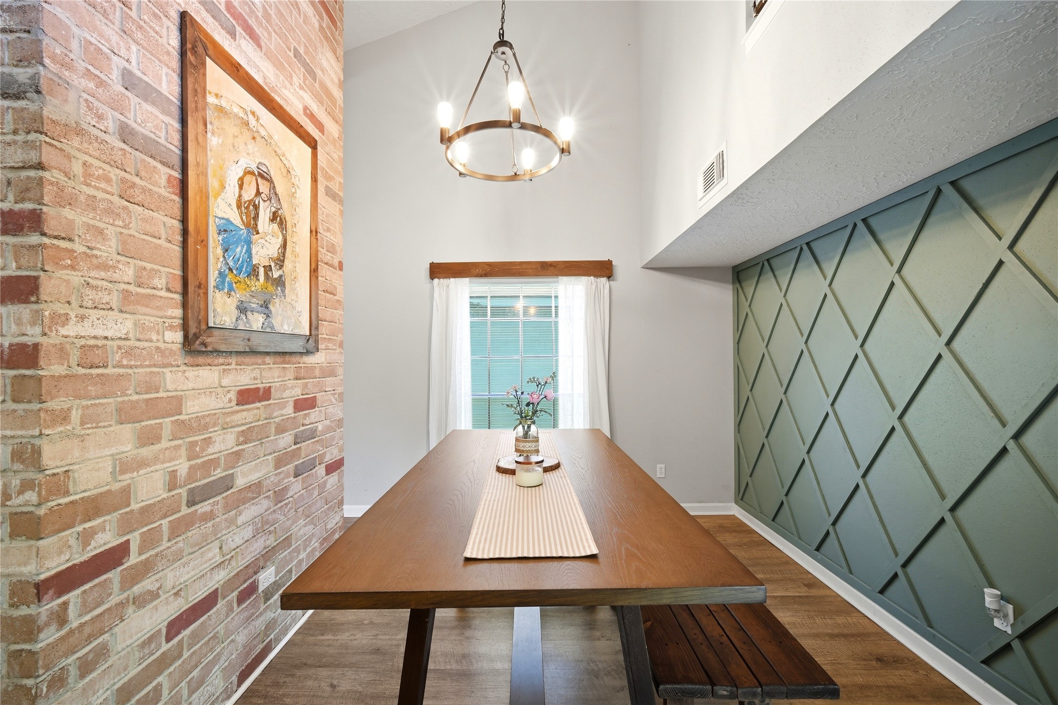 4718 Temple Bell Drive Spring, TX 77388 - Photo 14 of 50 This photo showcases a modern dining area with a high ceiling, featuring a stylish wood table and bench. The space is accented by a brick wall with artwork and a chic geometric accent wall. A large window with curtains brings in natural light, and a contemporary chandelier adds elegance.