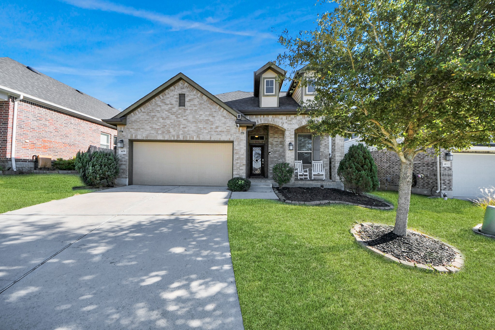 This beautiful home features a well-maintained brick exterior with a two-car garage, a manicured front lawn, and inviting front porch with seating.