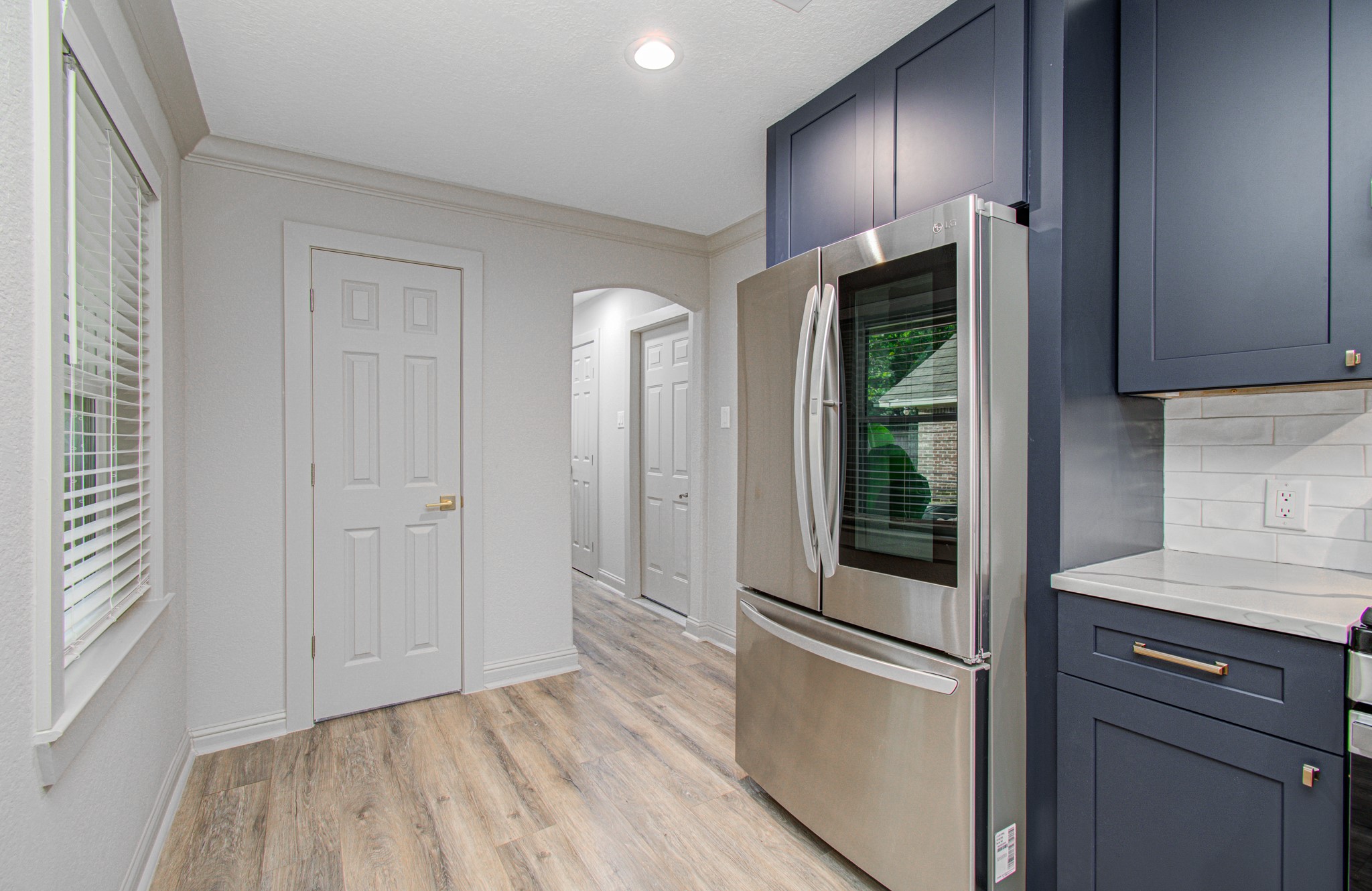 1801 Benbow Way Houston, TX 77080 - Photo 14 of 32 This kitchen features sleek navy cabinets, a modern stainless steel refrigerator, and light wood flooring. The space is brightened by natural light from a window, and a hallway leads to other parts of the home.