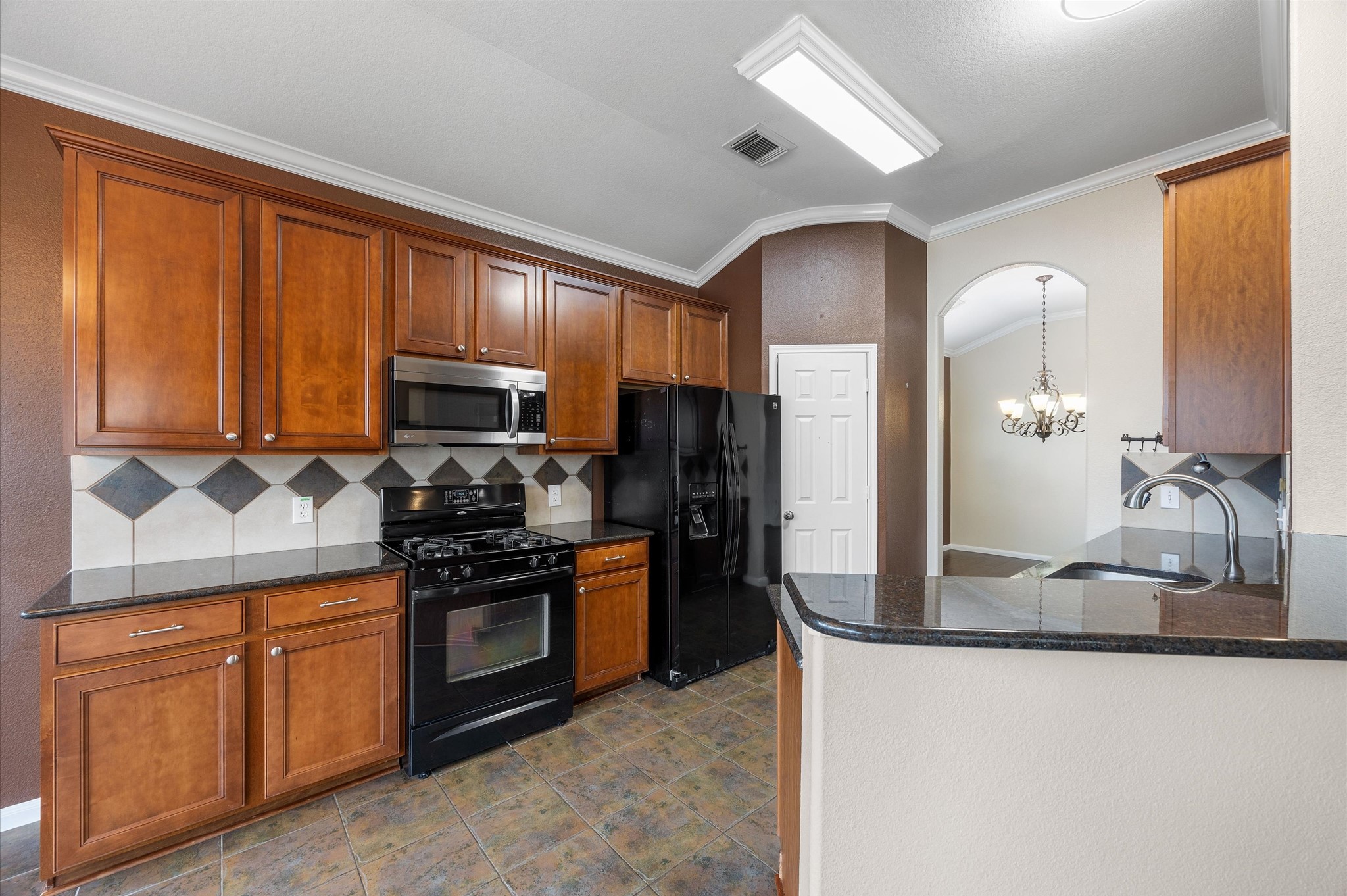 24538 Fort Settlement Drive Spring, TX 77373 - Photo 11 of 29 This kitchen features warm wooden cabinetry, a black appliance suite, and a stylish tile backsplash. It boasts a gas stove, built-in microwave, and a spacious countertop with an adjacent archway leading to a dining area with a chandelier.