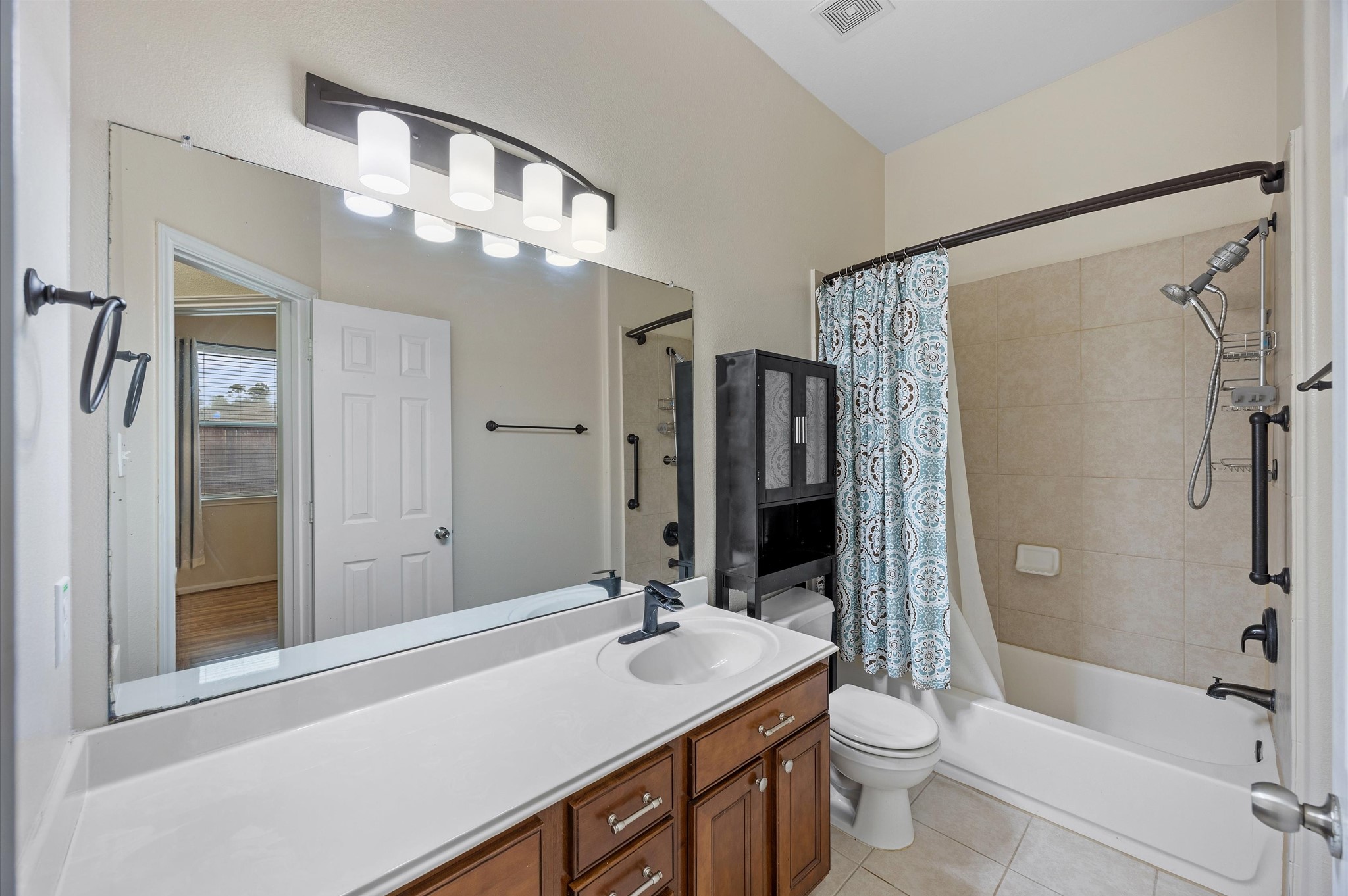 24538 Fort Settlement Drive Spring, TX 77373 - Photo 15 of 29 This photo shows a bright, modern bathroom featuring a single sink vanity with ample countertop space and wood cabinetry. The room includes a full bath with a tiled shower and a decorative curtain, enhanced by a large mirror and stylish lighting.