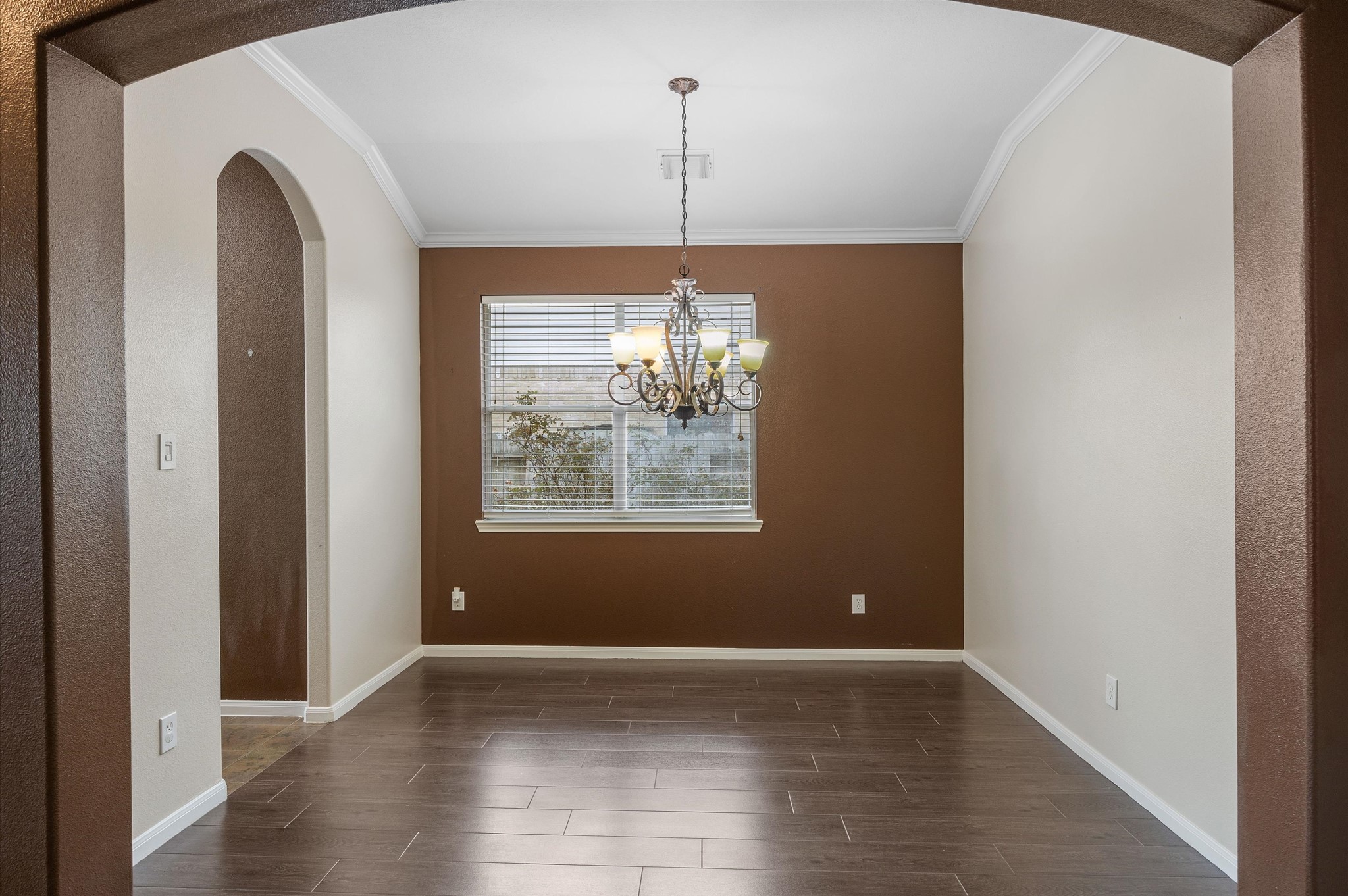 24538 Fort Settlement Drive Spring, TX 77373 - Photo 10 of 29 This photo showcases a dining room with a neutral color palette, featuring dark wood flooring and a two-toned wall design. A central chandelier provides elegant lighting, and a window brings in natural light. The space is framed by an arched doorway, adding a touch of architectural interest.