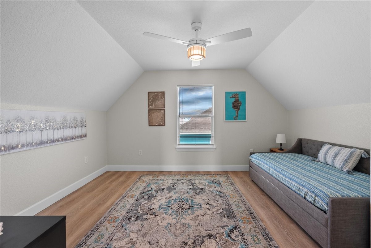 130 Beachcomber Avenue Surfside Beach, TX 77541 - Photo 19 of 33 This cozy room features a sloped ceiling with a ceiling fan, a single window, and light wood look flooring. It includes a daybed with striped bedding, a small bedside table with a lamp, and tasteful wall art, creating a welcoming and functional space.