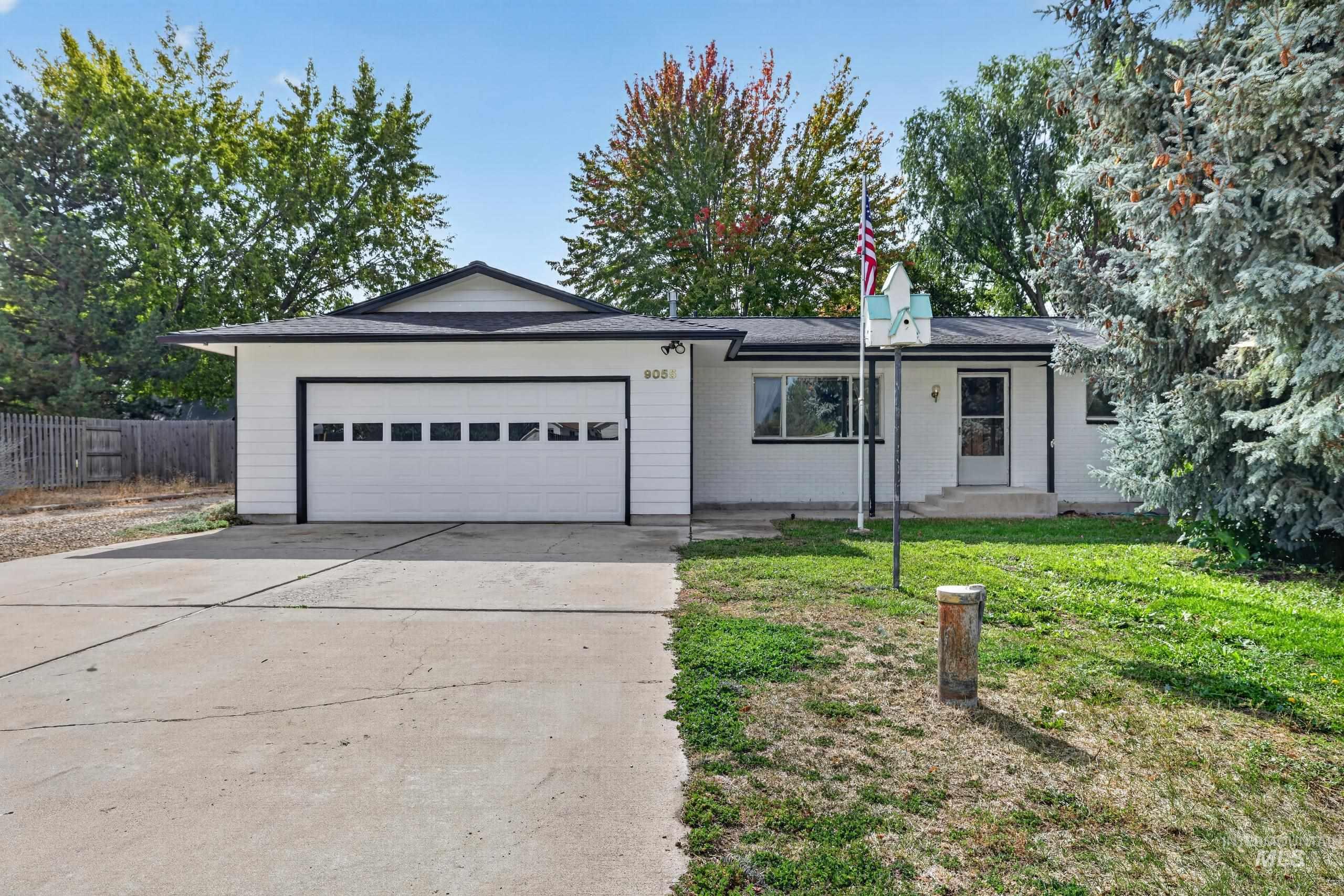 9055 West Highlander Road Boise, ID 83709 - Photo 39 of 39 White exterior siding with a black shingle roof, attached garage with white door, concrete driveway, and mature trees