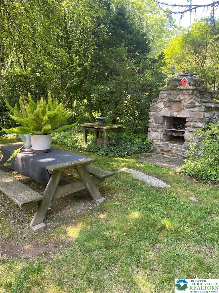 A rustic stone fireplace anchors this shaded outdoor living space surrounded by mature trees.
