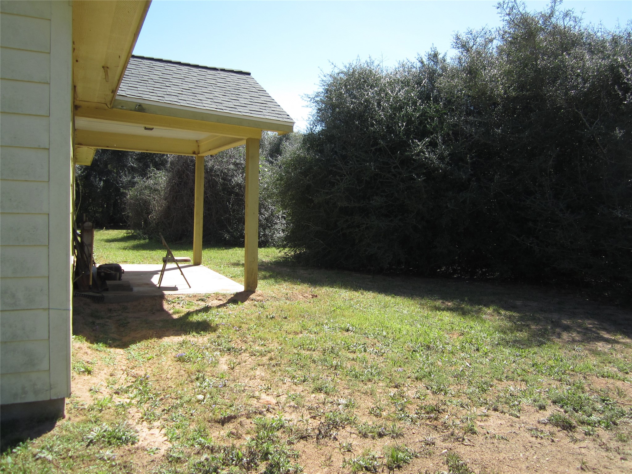 20697 Pierceall Road Hempstead, TX 77445 - Photo 31 of 35 This photo shows a small covered patio attached to the back of a house, with a grassy yard and dense shrubs providing privacy.