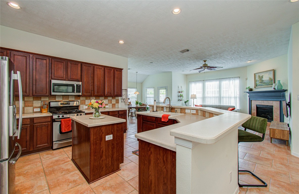 20007 Alcea Court Spring, TX 77379 - Photo 19 of 43 This spacious open-concept kitchen features rich wooden cabinetry, stainless steel appliances, and a central island. It flows seamlessly into a bright living area with large windows and a cozy fireplace, perfect for entertaining.