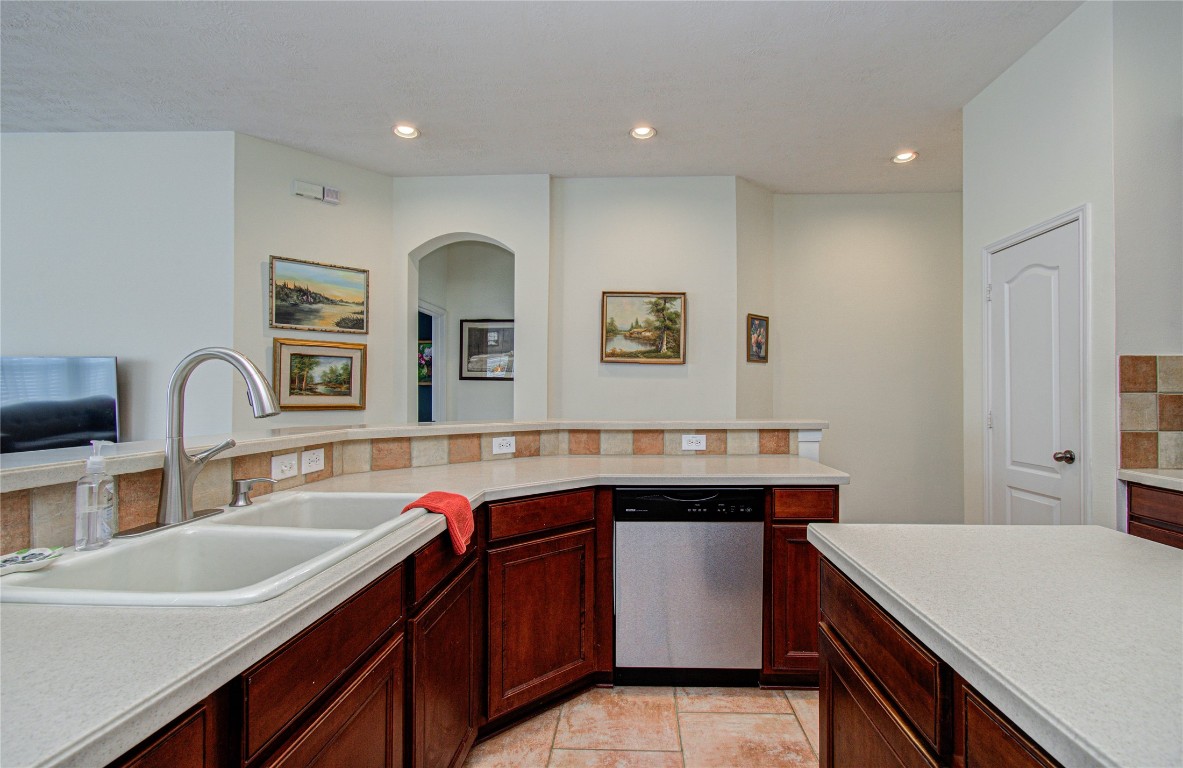 20007 Alcea Court Spring, TX 77379 - Photo 21 of 43 This kitchen features dark wood cabinetry, a double sink, and a stainless steel dishwasher. The countertops are light, and the space is accented with tile backsplash and recessed lighting. Art pieces add a touch of elegance to the open layout.