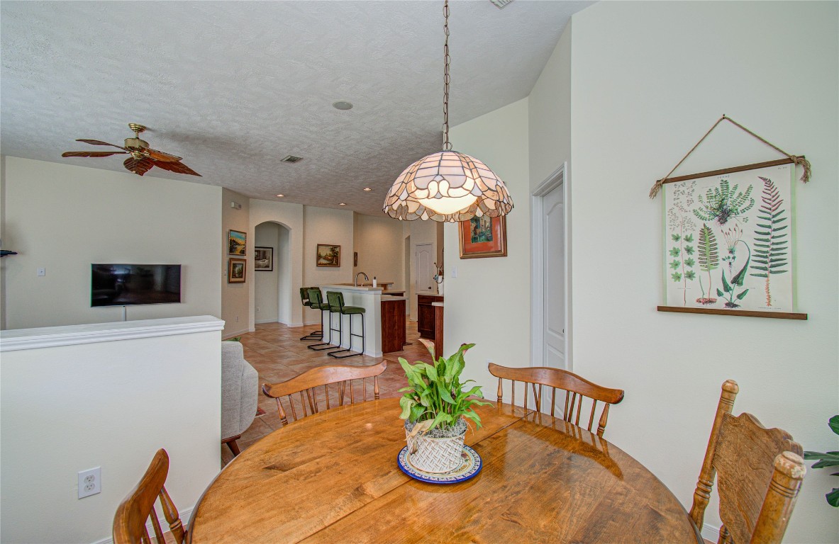 20007 Alcea Court Spring, TX 77379 - Photo 28 of 43 This cozy dining area features a wooden table with chairs, a decorative hanging light, and a wall with botanical art. It opens into a living space with a ceiling fan and a wall-mounted TV, leading to a kitchen with bar seating, creating a warm, inviting atmosphere.