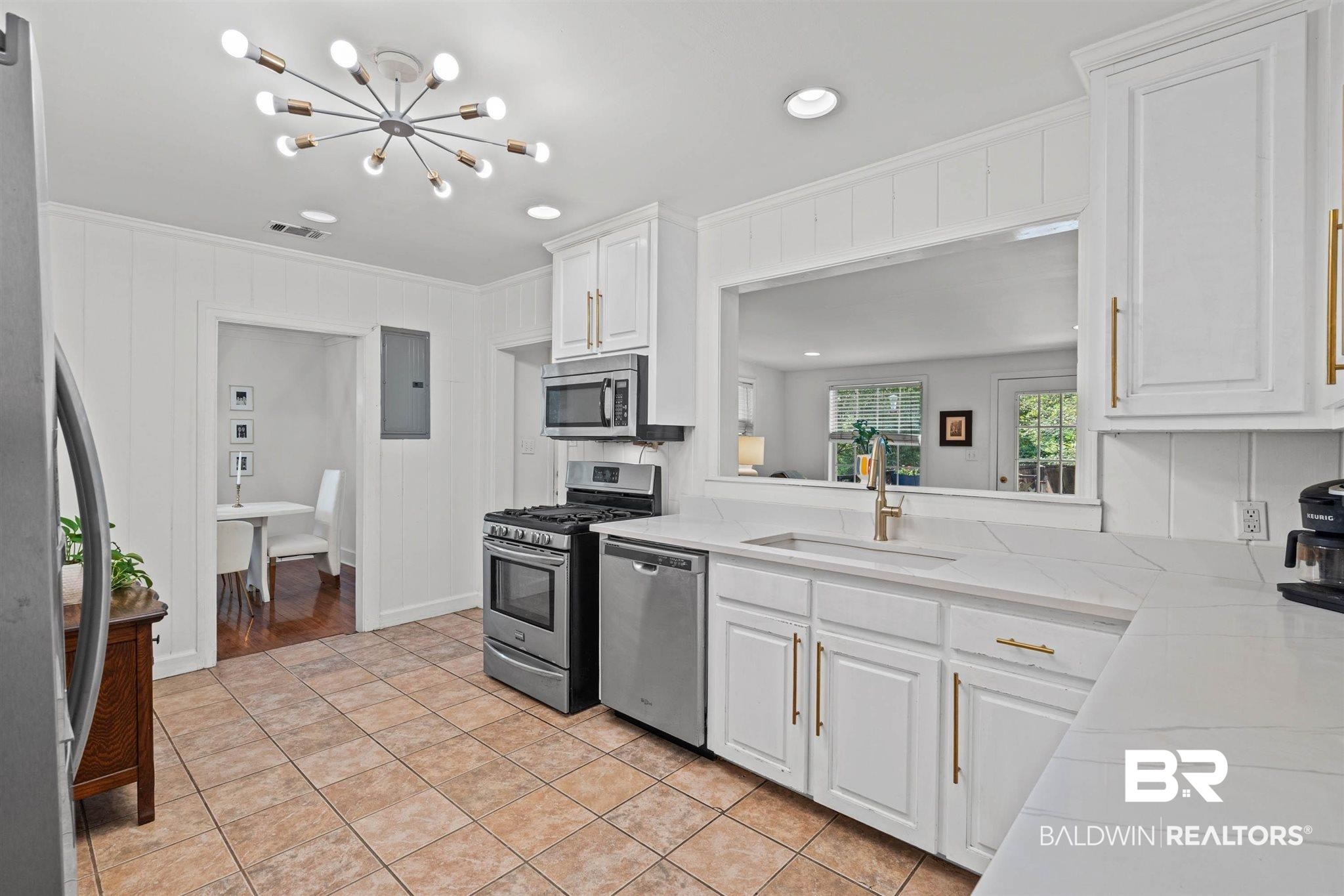 3751 Old Shell Road Mobile, AL 36608 - Photo 9 of 21 White kitchen featuring a pass-through window, stainless steel appliances, shaker-style cabinetry with gold-finish hardware, quartz countertops, and a modern sputnik-style light fixture