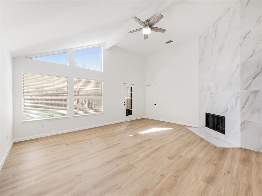 Vaulted ceiling with exposed beam, wood-finish flooring, white marble-look fireplace, large picture windows, and a ceiling fan
