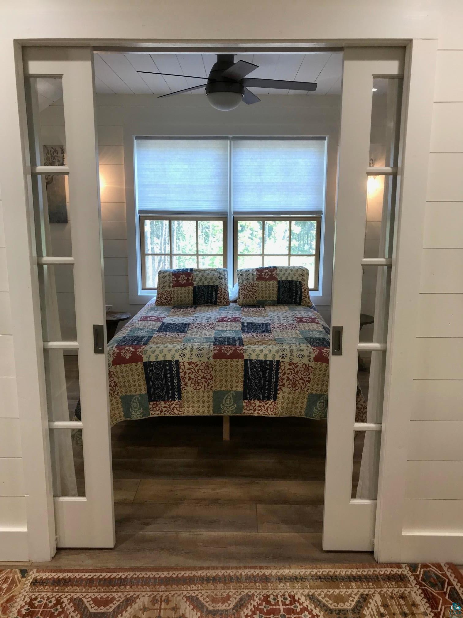 70745 Range Line Road Iron River, WI 54847 - Photo 30 of 40 Sliding barn doors with transom windows, wood-finish flooring, two large windows, a ceiling fan with light, and shiplap wall paneling