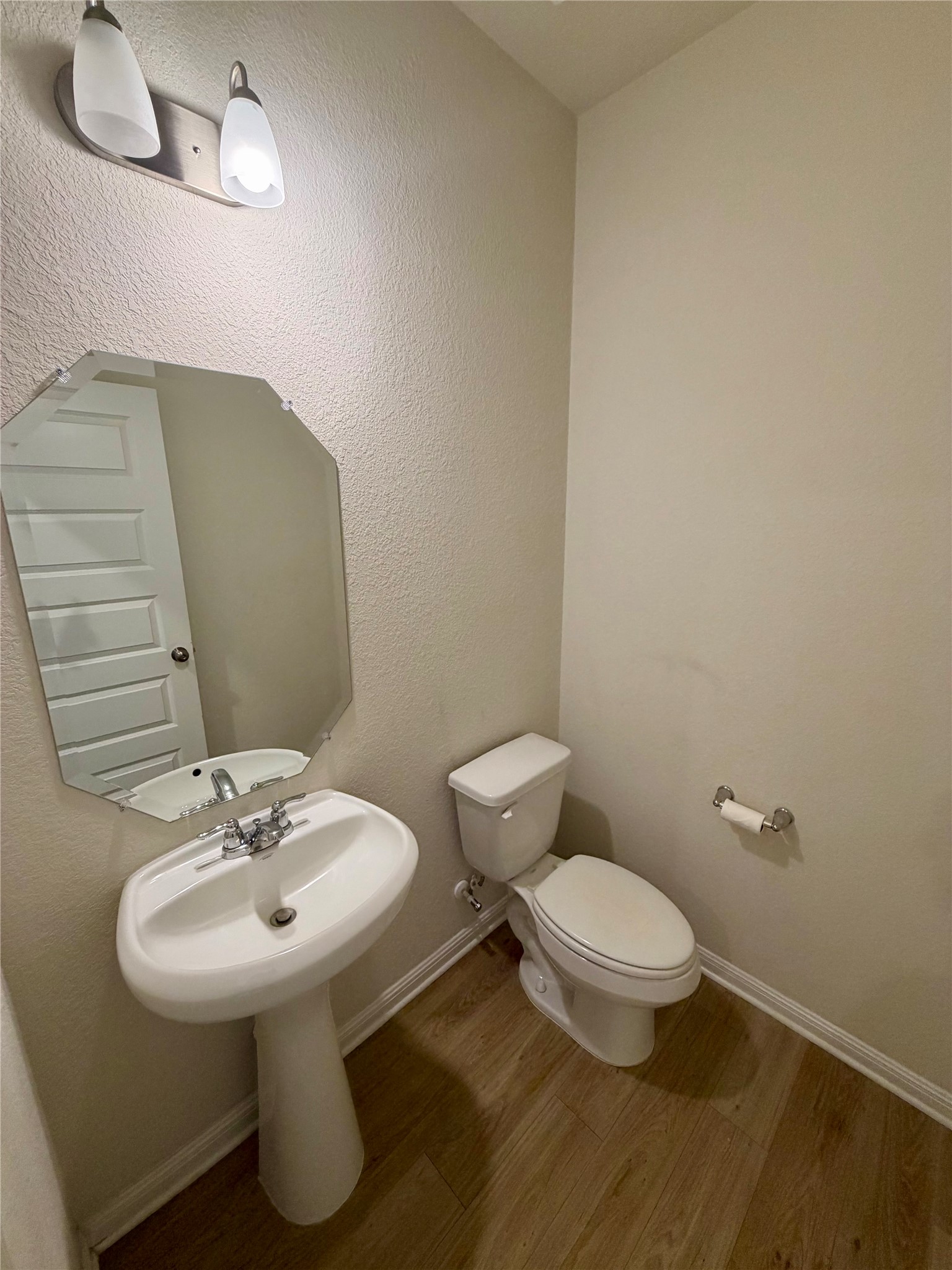305 Tolo Drive Elgin, TX 78621 - Photo 12 of 27 Powder room featuring a white pedestal sink with chrome fixtures, an octagonal mirror, a toilet, and wood-finish flooring