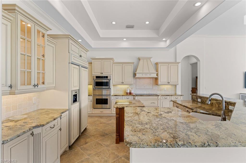 7506 Hogan Court Naples, FL 34113 - Photo 34 of 49 Kitchen featuring cream cabinetry, backsplash, arched walkways, light stone counters, and a raised ceiling