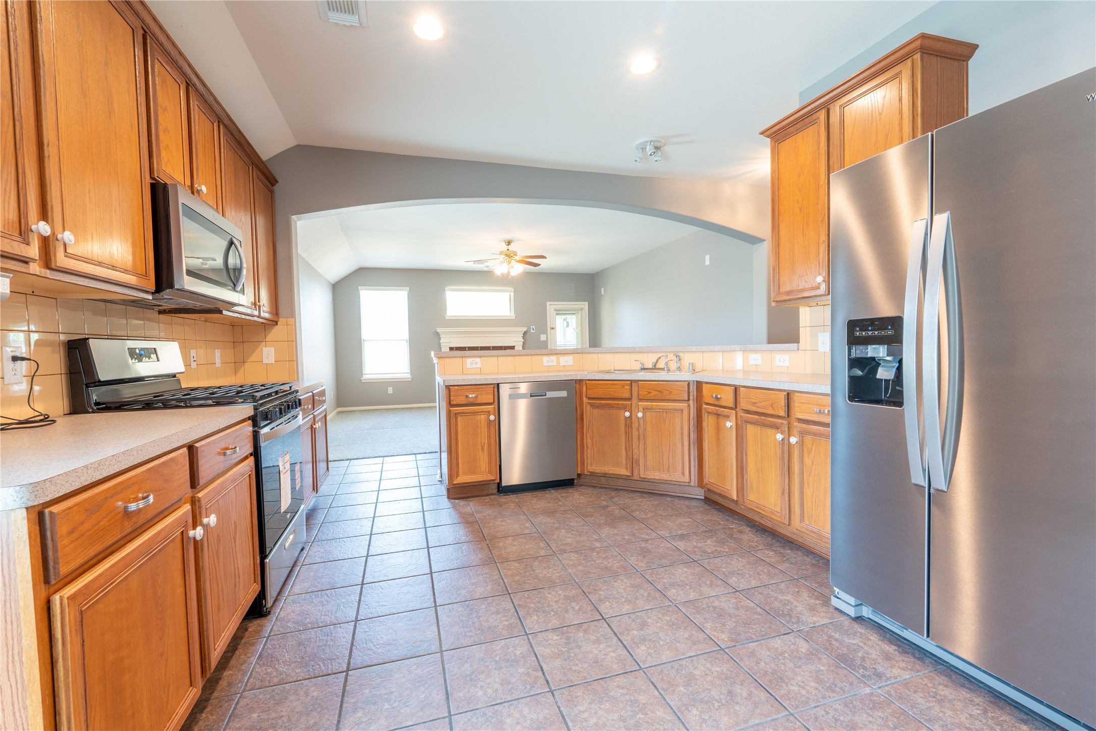 10619 Crownsedge Drive Spring, TX 77379 - Photo 3 of 38 Spacious kitchen with stainless steel appliances, wooden cabinetry, and tile flooring. Open layout leads to a bright living area with ceiling fan and natural light.