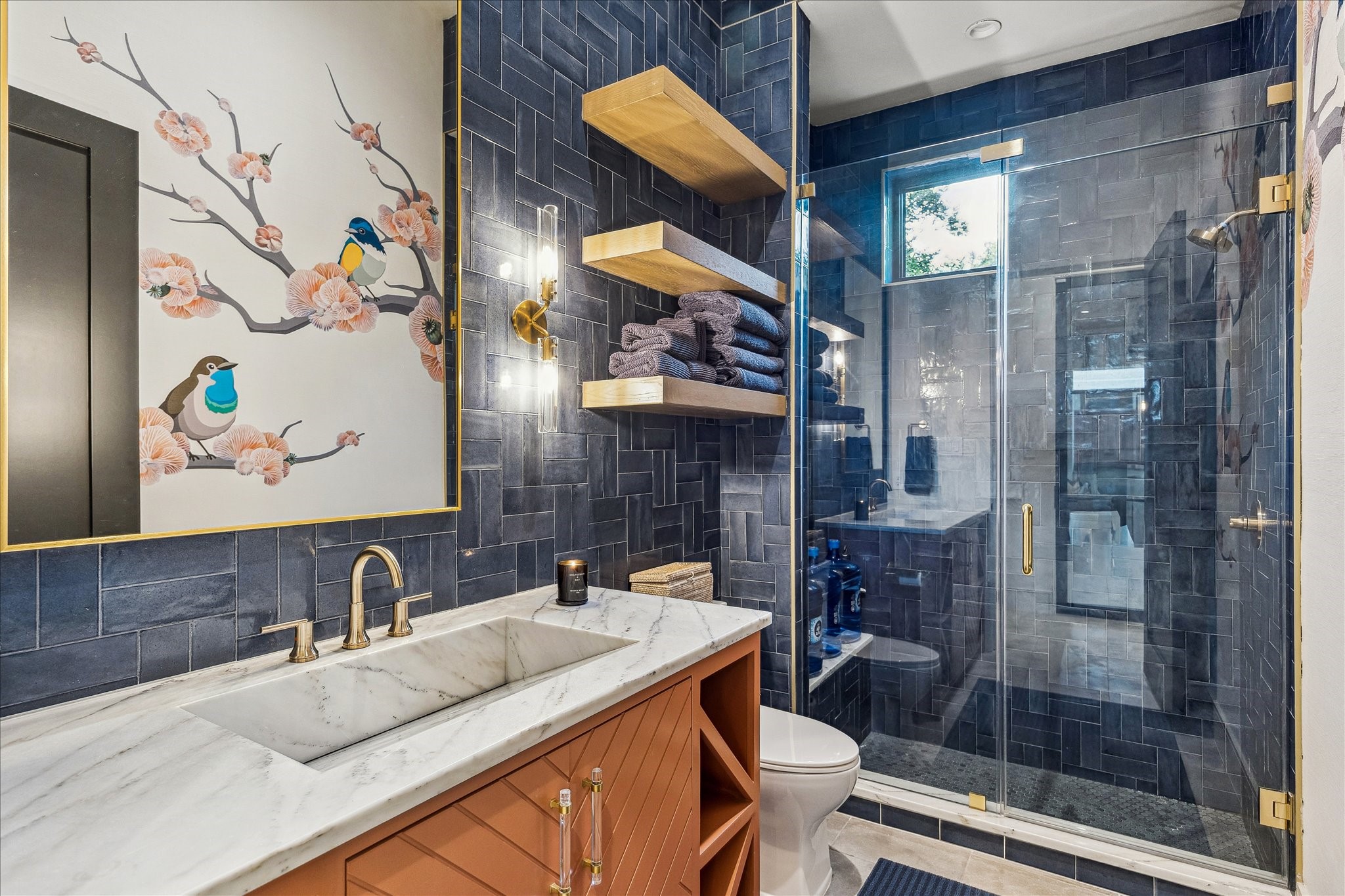 3807 Westerman Street Houston, TX 77005 - Photo 20 of 50 The first-floor full bath features acrylic and brass hardware, a marble integrated sink with storage cabinets below, and elegant vanity sconces. A walk-in shower with seamless glass door and designer tile backsplash adds a luxurious touch. Floating white oak shelves and designer wallpaper complete the space, combining style and functionality.
