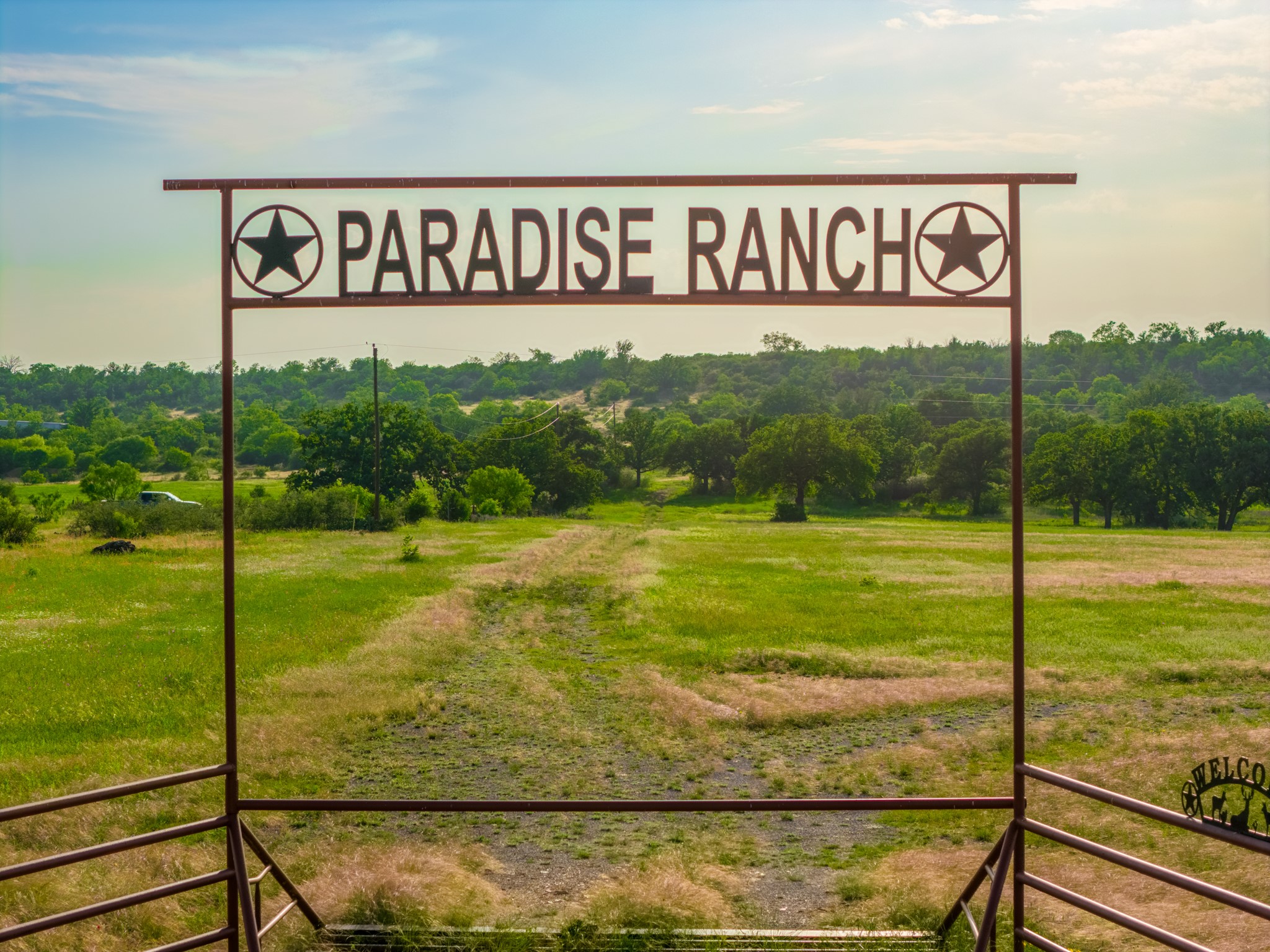 11079 Cr 266 Rochelle Tx 76872 Rochelle, TX 76872 - Photo 26 of 40 Custom metal entry gate featuring "Paradise Ranch" lettering and star accents, framed by expansive green fields and mature trees under a clear sky