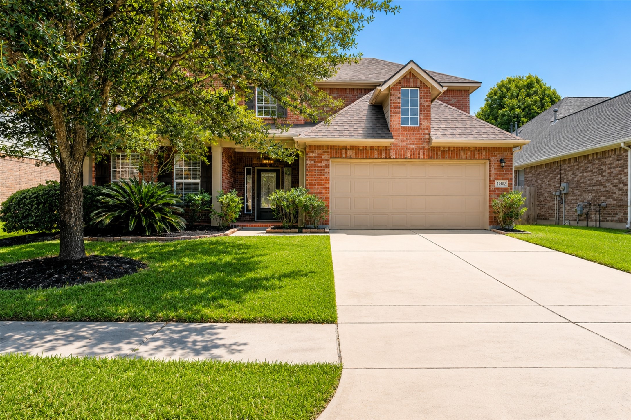 17402 Rainer Valley Lane Humble, TX 77346 - Photo 1 of 33 This photo showcases a charming brick home with a three-car tandem garage, a well-maintained front lawn, and a mature tree offering shade. The entrance is inviting, featuring a covered porch with decorative plants.