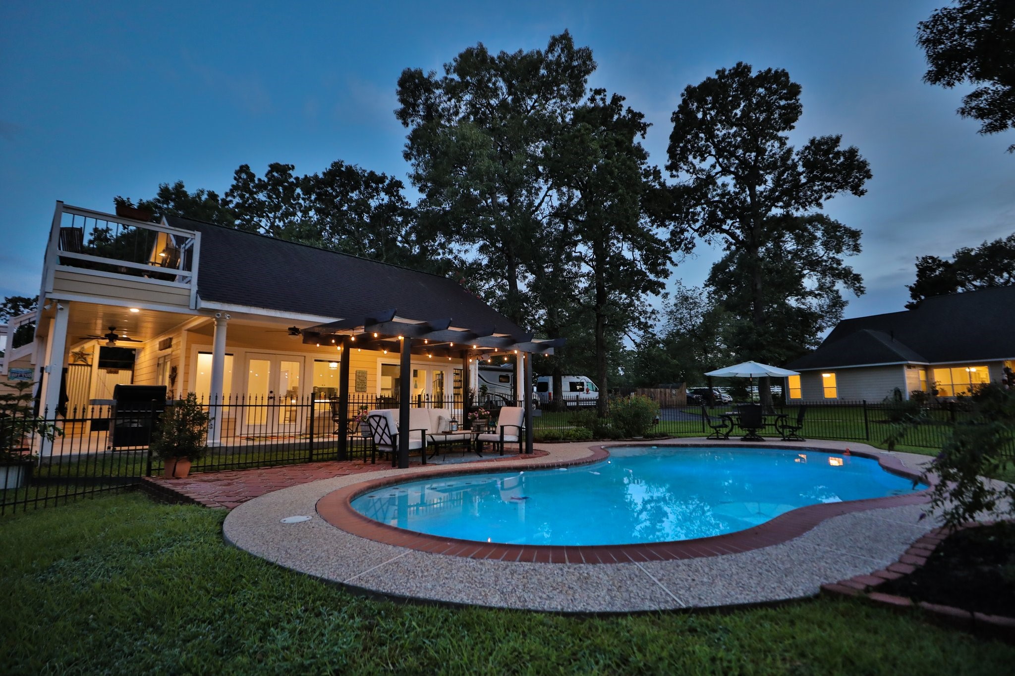 34310 High Point Drive Magnolia, TX 77355 - Photo 38 of 48 This photo showcases a charming backyard featuring a well-lit pool with surrounding patio space and guest house. The adjacent deck and seating area under a pergola offer a perfect spot for relaxation or entertaining. Lush greenery and mature trees provide a peaceful, private atmosphere, complemented by the warm glow from the home's exterior lighting.