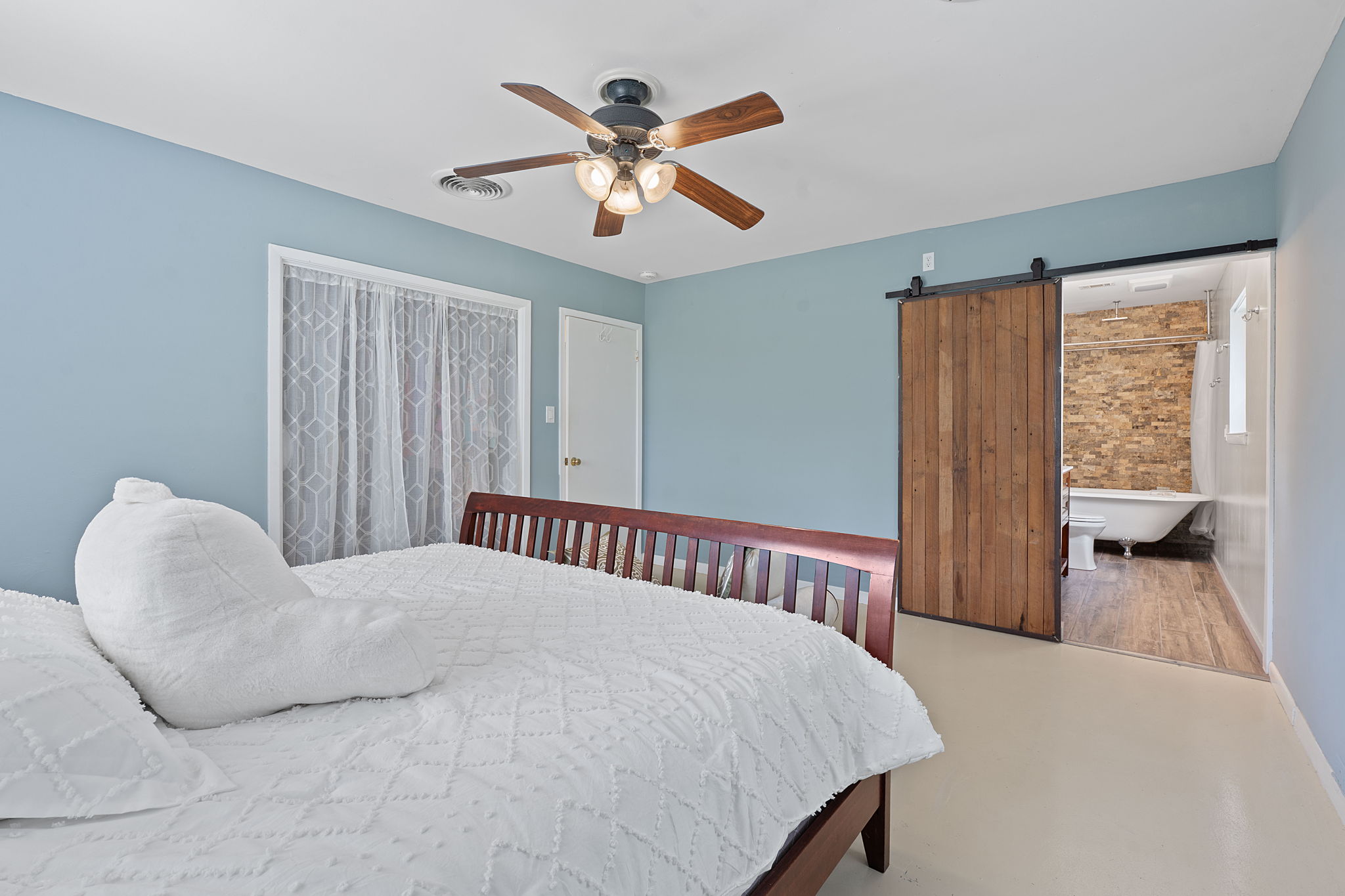8705 South View Road Austin, TX 78737 - Photo 16 of 39 Serene primary bedroom with soft blue walls, wooden ceiling fan, and sliding barn door to ensuite bathroom with clawfoot tub