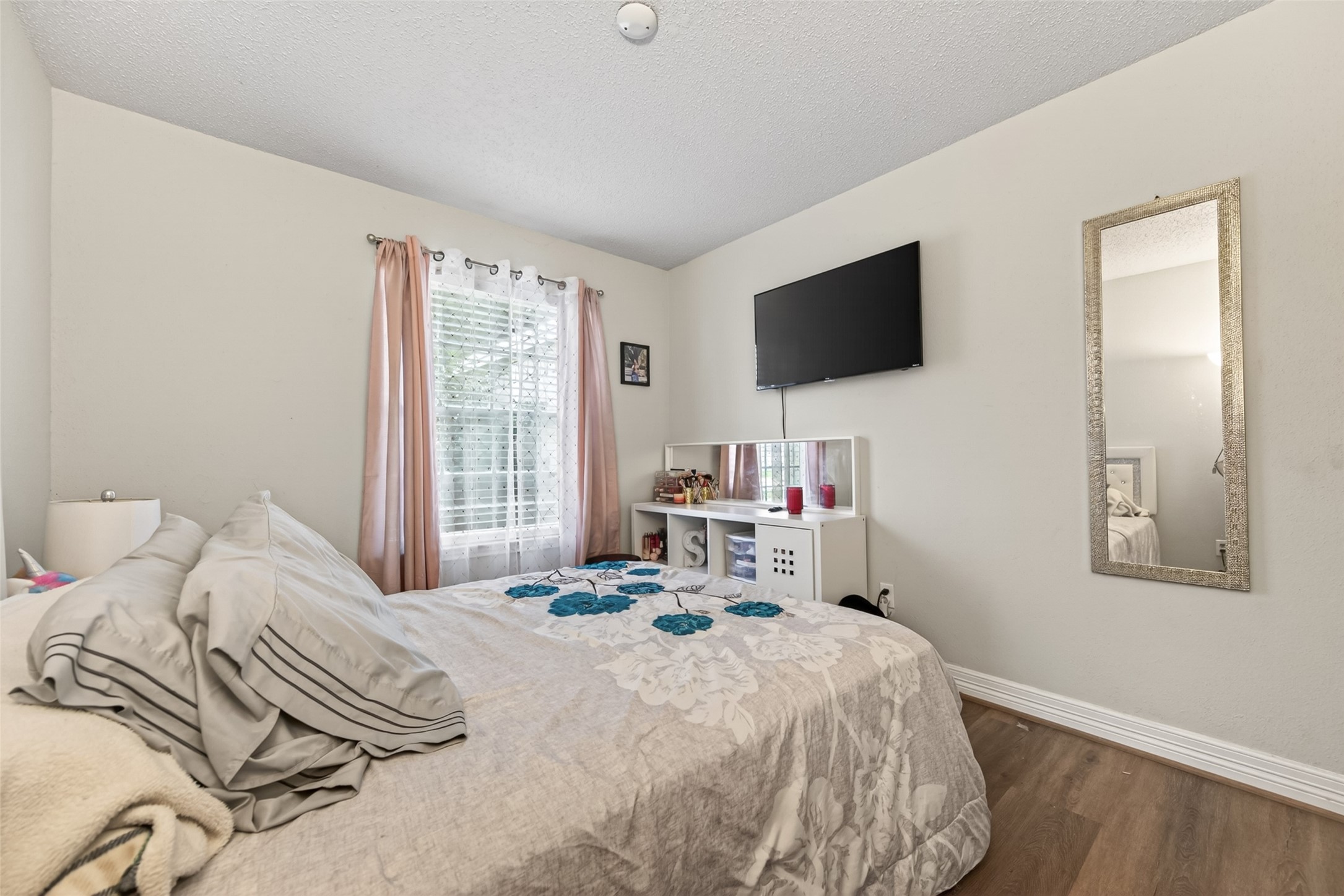 18446 North Willow Bluff Road Katy, TX 77449 - Photo 28 of 33 Cozy bedroom with a large window providing natural light, featuring a mounted TV, a decorative mirror, and wood flooring for a warm, inviting feel.