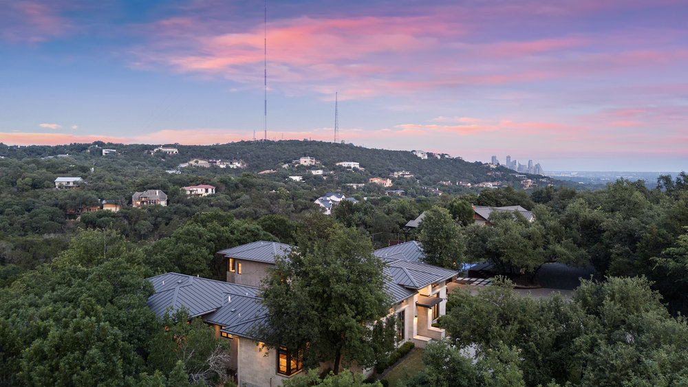 1905 Paisano Road Austin, TX 78746 - Photo 37 of 40 An aerial perspective reveals the tranquil hillside setting of a residence amidst lush trees, with distant city views under a twilight sky.