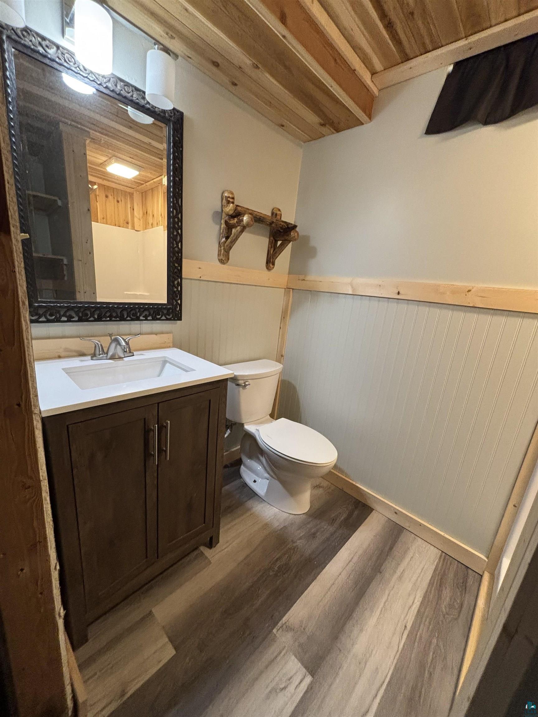 444 Fernberg Road, Unit 11 Ely, MN 55731 - Photo 17 of 19 Bathroom featuring a dark wood-finish vanity with an integrated sink and chrome faucet, wood-finish flooring, a white toilet, and beadboard paneling