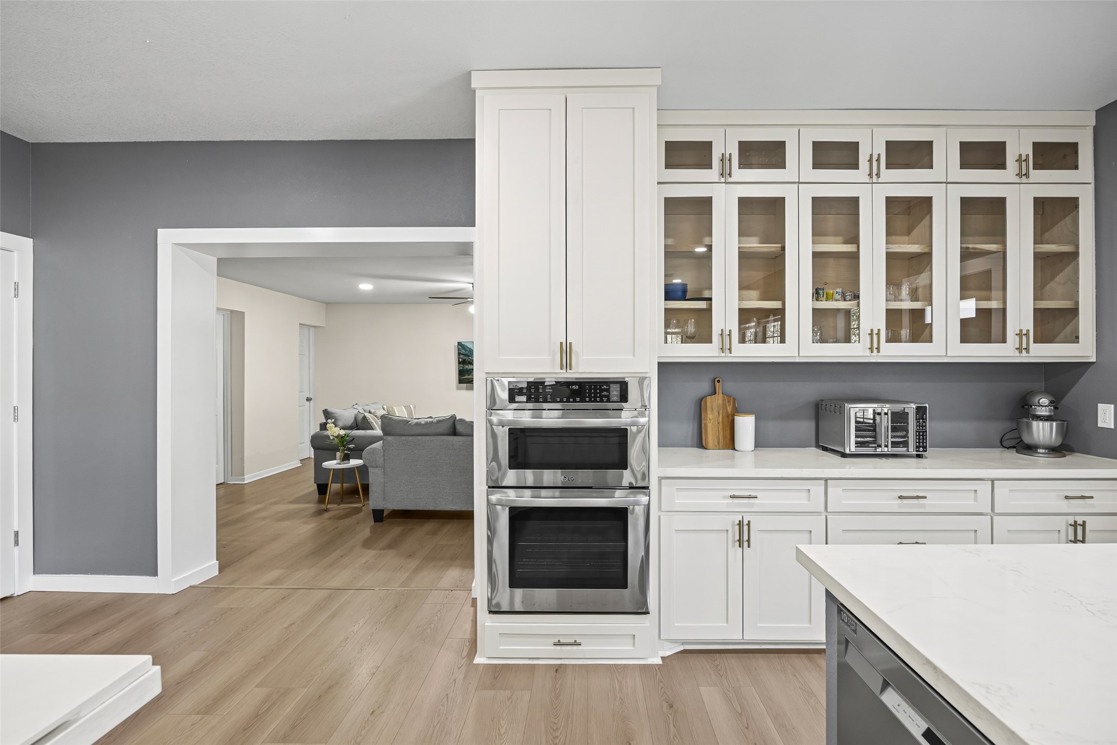 15080 Usher Lane Splendora, TX 77372 - Photo 20 of 44 This photo showcases a modern kitchen with sleek white cabinetry, glass-front upper cabinets, and stainless steel appliances. The open layout connects to a cozy living area with wooden floors, creating a seamless flow between spaces.