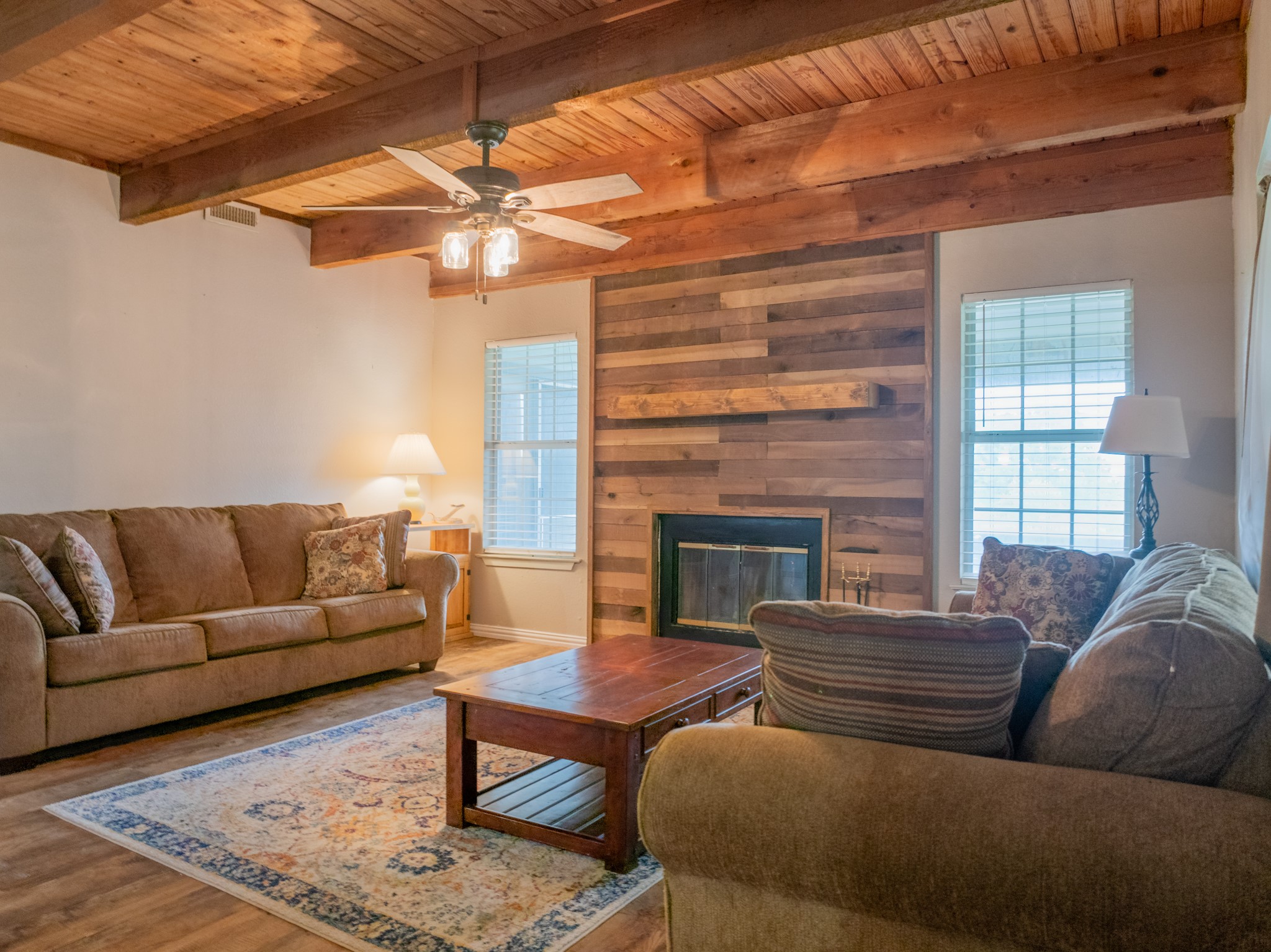 11079 Cr 266 Rochelle Tx 76872 Rochelle, TX 76872 - Photo 13 of 40 Living area featuring a wood-planked ceiling with exposed beams, a ceiling fan with integrated lighting, wood-finish flooring, a fireplace with a wood-plank accent wall, and multiple windows