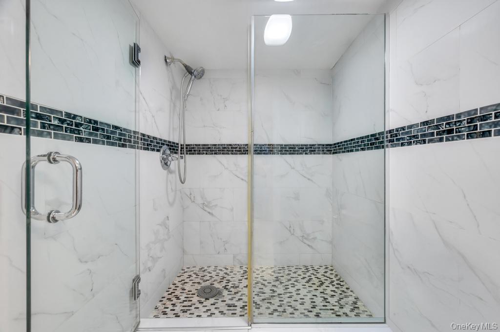 89-13 84th Street Queens, NY 11421 - Photo 28 of 34 Glass-enclosed shower featuring white wall tiling with dark accent band, pebble-style floor tiling, and chrome fixtures