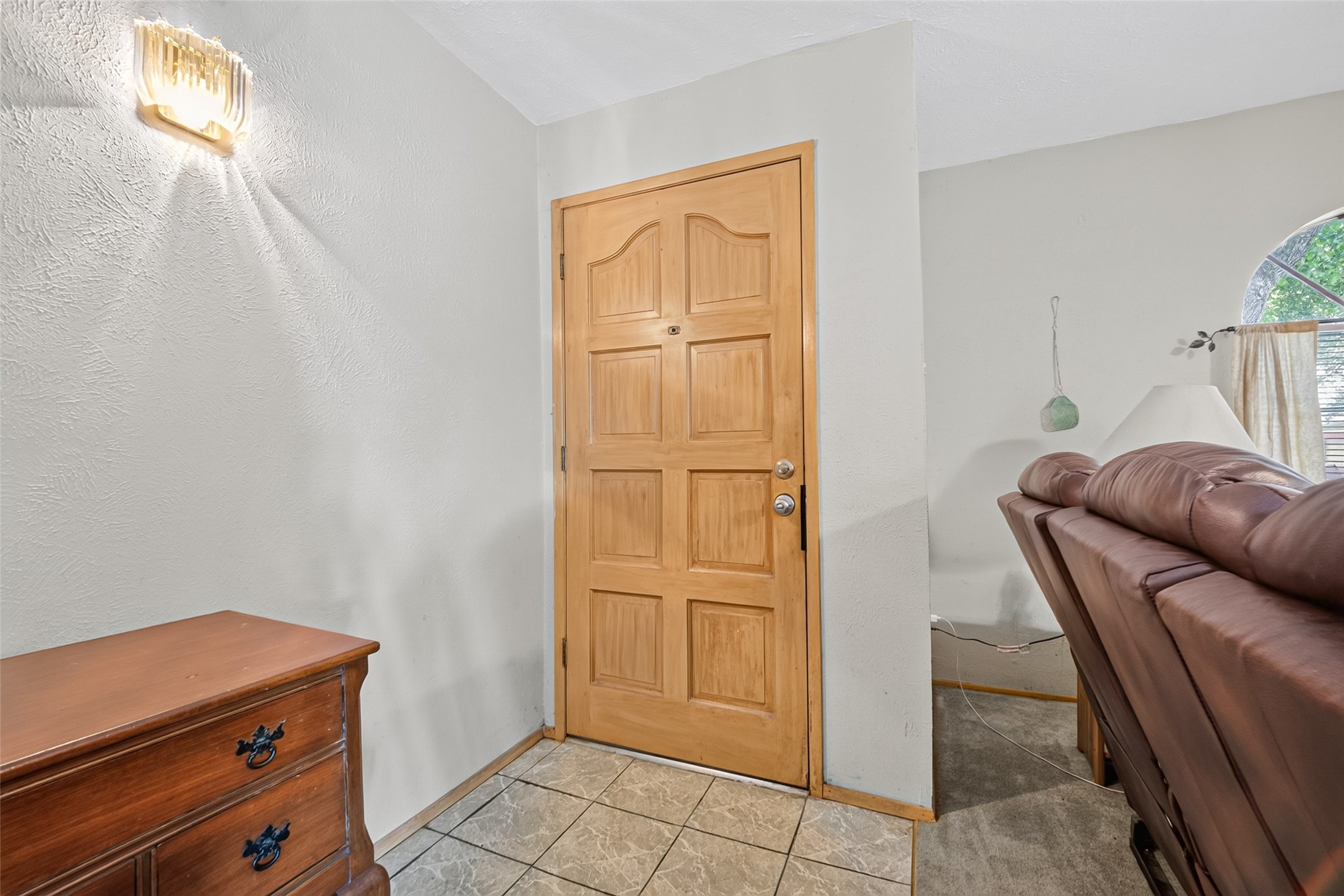 22926 Millgate Drive Spring, TX 77373 - Photo 5 of 34 This photo shows a cozy entryway with a wooden front door, tiled flooring, and a small chest of drawers. To the right, there's a glimpse of a living room with brown leather seating and a window with curtains.