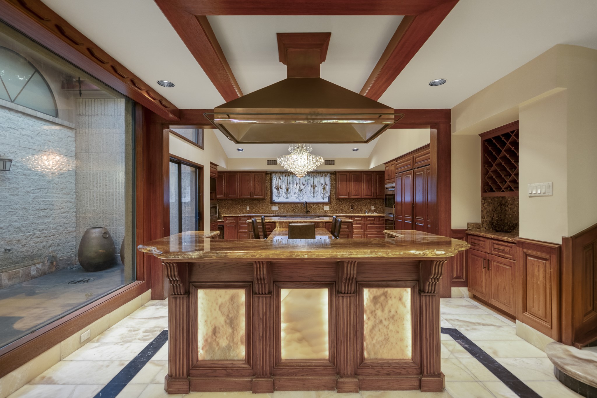 6535 Buffalo Speedway Houston, TX 77005 - Photo 10 of 34 Part of the expansive main kitchen, this bar-height island features a built-in cooktop and beautifully backlit stone that glows with warmth and elegance. Rich wood cabinetry and generous counter space make it ideal for both casual dining and everyday cooking.