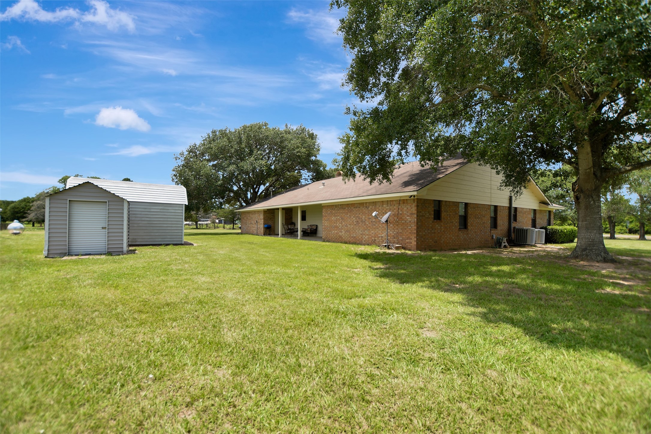 341 South S Loop Bellville, TX 77418 - Photo 30 of 33 This photo shows a spacious backyard featuring a brick house with a sloped roof and a shed. The yard is grassy with mature trees providing shade, creating a peaceful and private outdoor space.