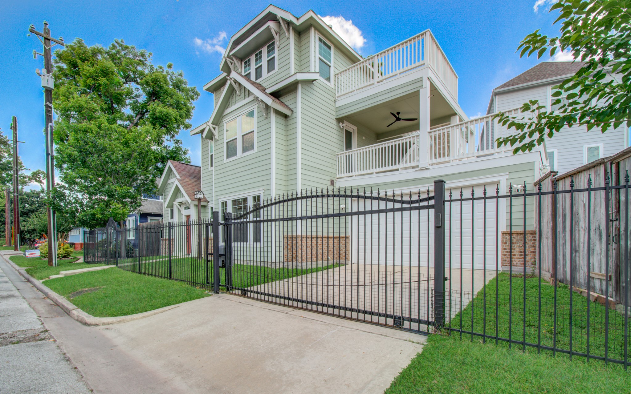 6621 North Main Street Houston, TX 77009 - Photo 3 of 41 This photo showcases a modern, multi-story home with a light green exterior and white trim. It features a spacious balcony, a large garage, private automatic driveway gate, and is secured by a stylish black metal fence. The property is well-maintained with a neatly manicured lawn and is situated on a quiet street.