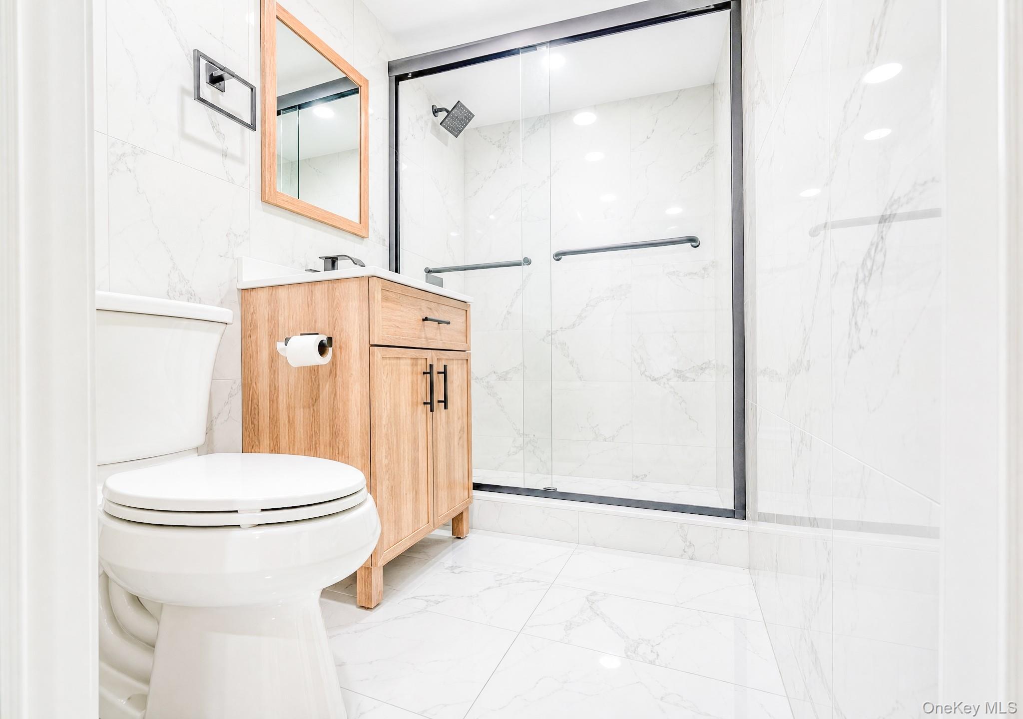 61 Cheshire Road Bethpage, NY 11714 - Photo 20 of 25 Bathroom featuring a frameless glass shower enclosure with matte black trim, a wood-finish vanity with ample storage, a rectangular wood-framed mirror, and white tile flooring and walls