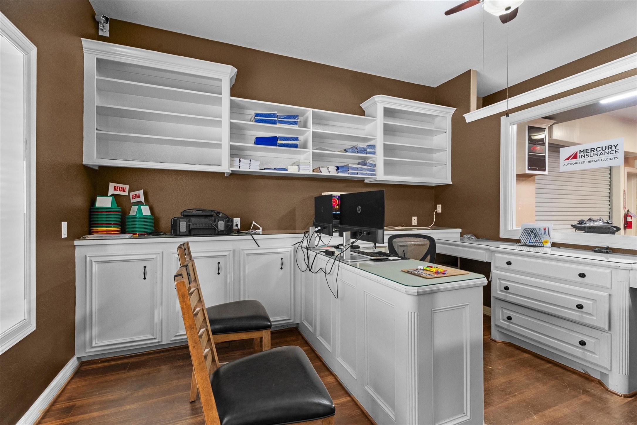 11066 Sh 242 Conroe, TX 77385 - Photo 14 of 21 This photo shows a home office with white cabinetry and shelving, featuring a built-in desk area. The room has a warm brown wall color, hardwood flooring, and a ceiling fan. It's well-lit and includes ample storage space.