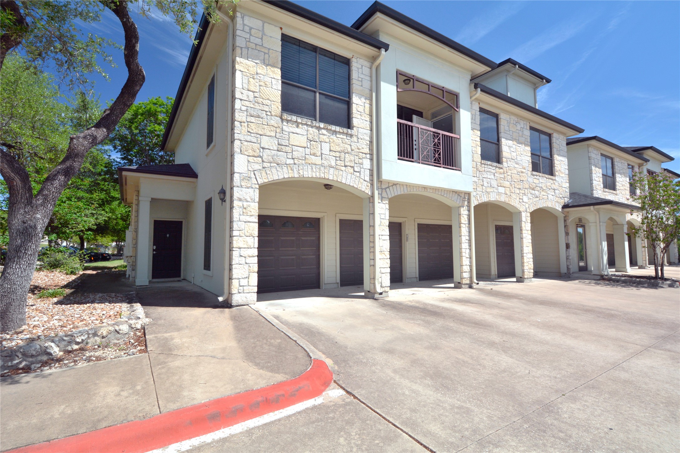 Multi-story property featuring a stone and stucco facade, arched garage entrances, dark garage doors, and an upper-level balcony with decorative railing