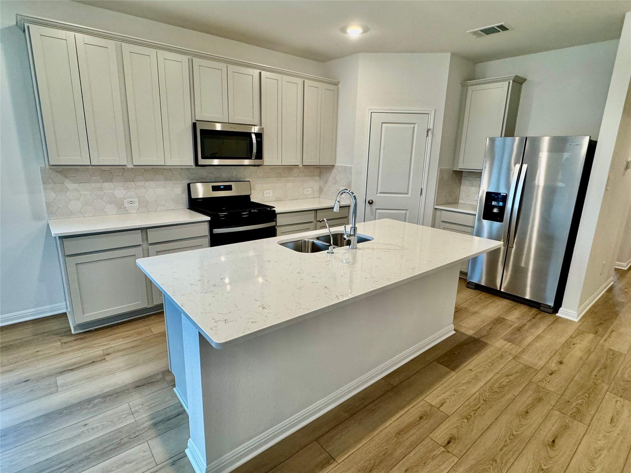 Undisclosed Address Kyle, TX 78640 - Photo 2 of 17 Kitchen featuring appliances with stainless steel finishes, light stone counters, a center island with sink, backsplash, and light wood-type flooring
