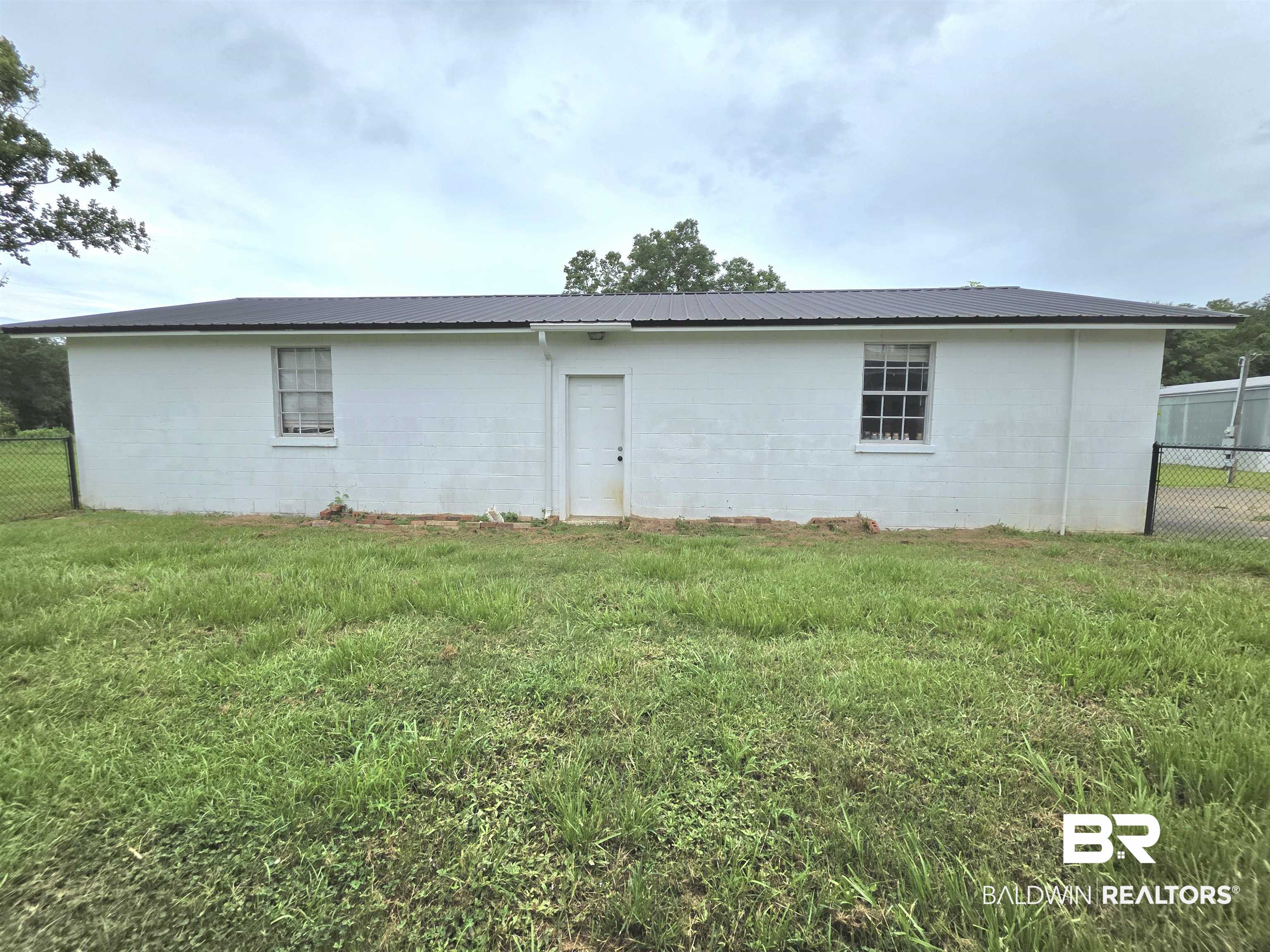 558 Stanton Road Atmore, AL 36502 - Photo 20 of 29 White masonry exterior with a dark metal roof, featuring a single entry door, two windows with grilles, and a chain-link fence