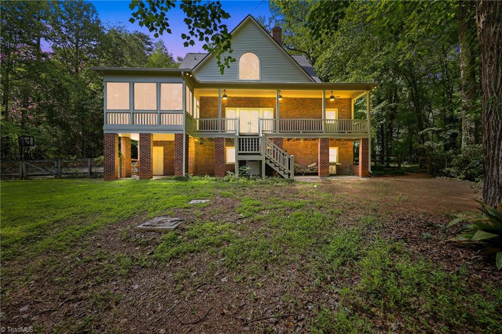 8861 Boylston Road Colfax, NC 27235 - Photo 38 of 50 The backyard is designed for both relaxation and functionality. Enjoy the expansive covered porch and screened-in porch overlooking a private, wooded setting, while the walk-out basement offers additional covered outdoor living space and a separate entrance—ideal for guests or multi-generational living. Surrounded by mature trees, this peaceful retreat offers the perfect balance of privacy, space, and everyday enjoyment. (Enhanced Twilight Rear Elevation)