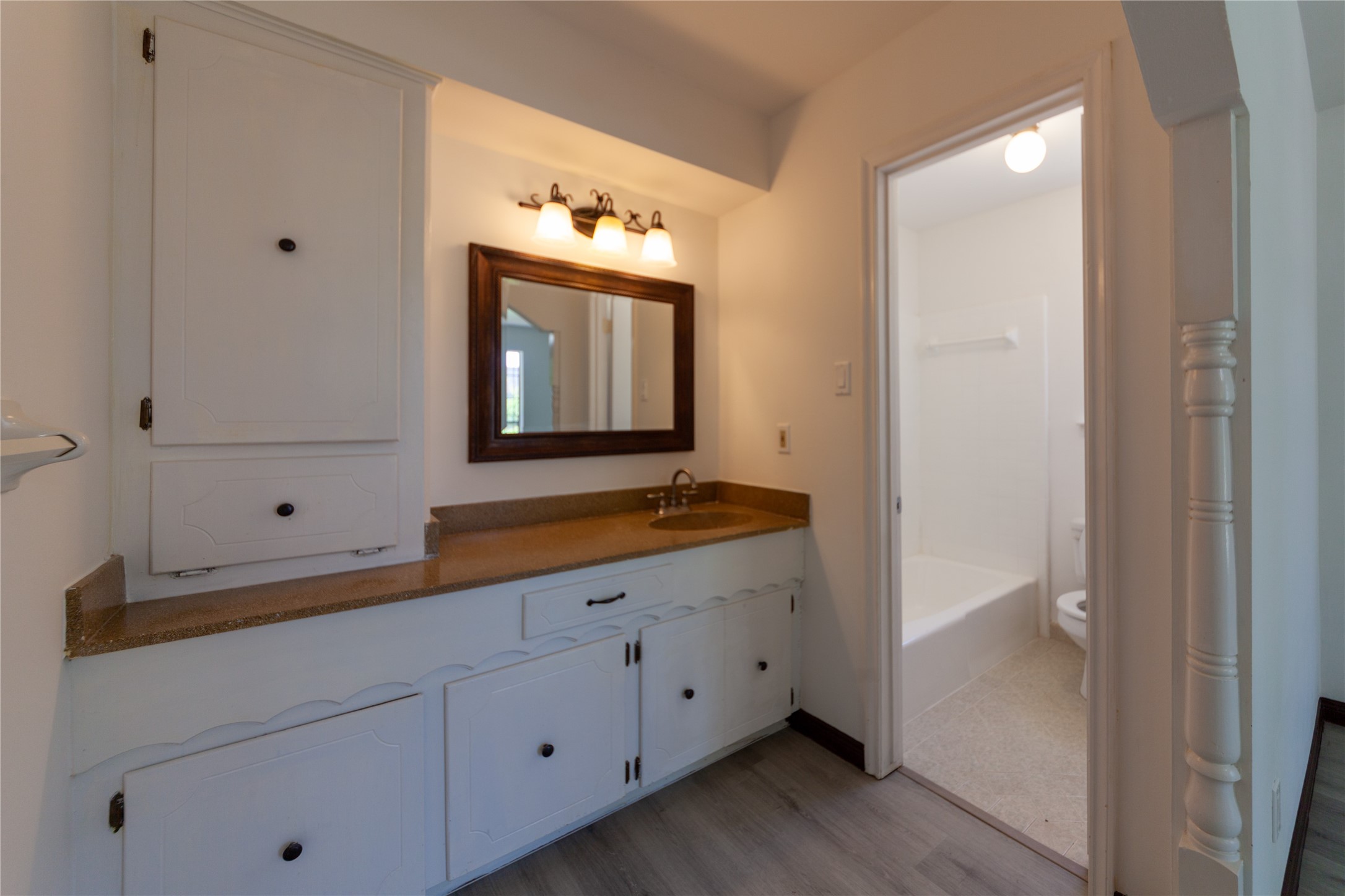 8826 Roos Road Houston, TX 77036 - Photo 16 of 42 A primary bathroom with a classic vanity featuring ample cabinet storage, a large mirror, and overhead lighting. Adjacent is a room with a bathtub and toilet, offering a functional layout with a clean, bright aesthetic.