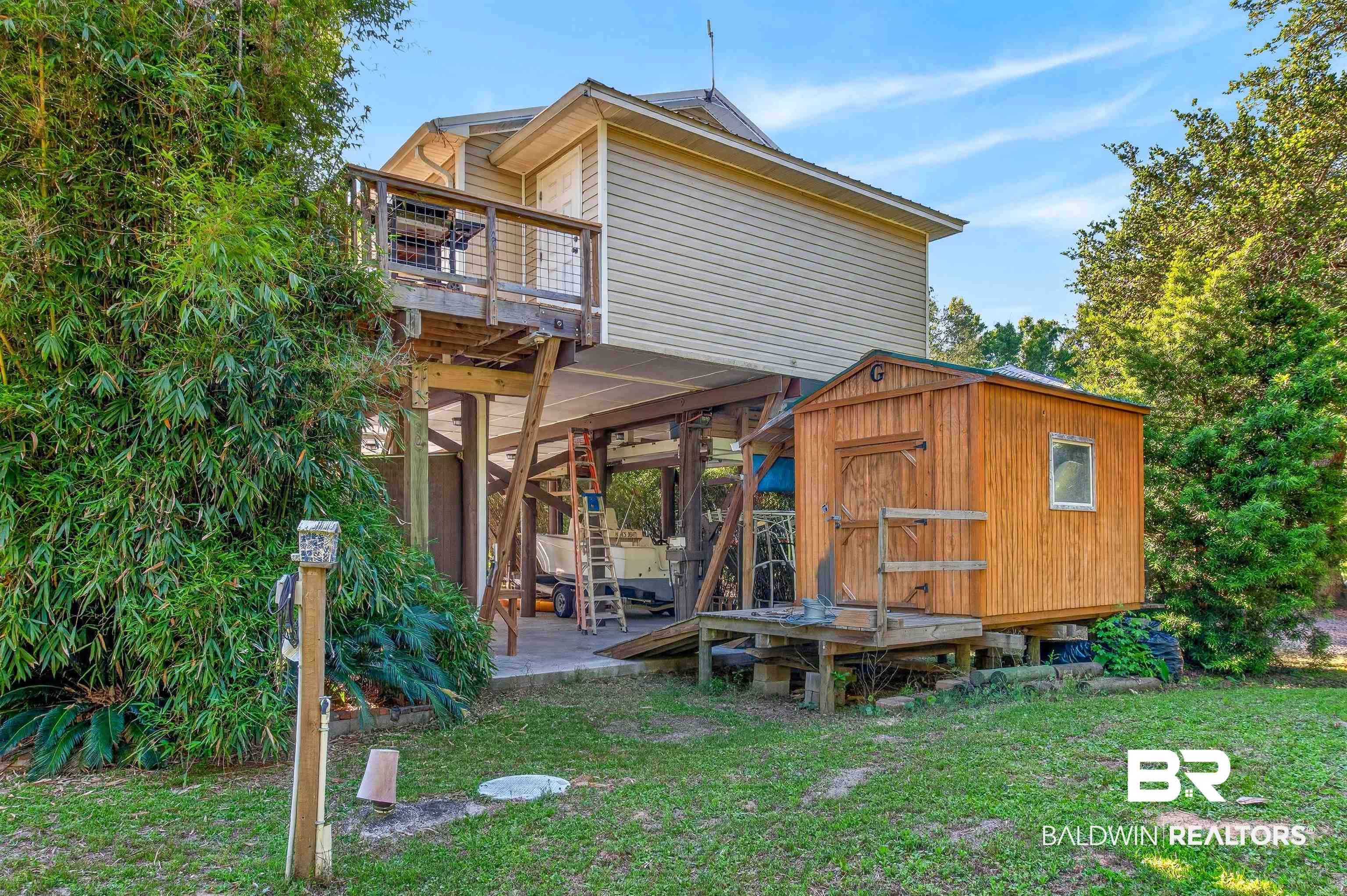 9624 North Bay Street Foley, AL 36535 - Photo 49 of 59 Elevated residence featuring a second-story deck with wood railing, a ground-level covered area supported by pilings, a detached wood-paneled shed with a window, and surrounding mature landscaping