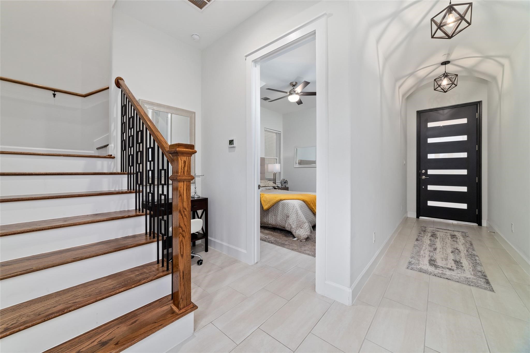 710 Delano Street, Unit A Houston, TX 77003 - Photo 15 of 32 This modern entryway with sleek tile flooring and contemporary lighting fixtures. A stylish staircase with wooden steps and a black railing leads to the upper level. The hallway opens to a room on the right, showcasing a cozy bedroom with a ceiling fan. The front door has a chic, horizontal panel design, adding to the home's modern aesthetic.