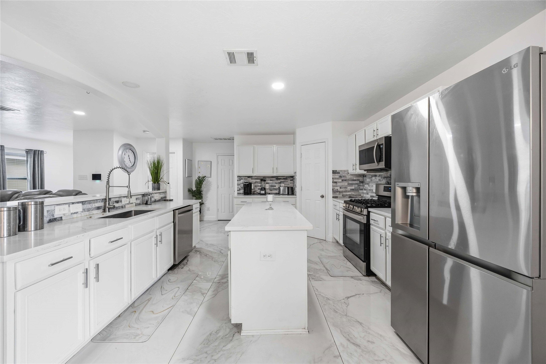 Bright, modern kitchen featuring a functional galley layout with a large center island, crisp white cabinetry, and expansive marble-look tile flooring. Stainless steel appliances, including a refrigerator and range, pair seamlessly with sleek countertops and a contemporary tile backsplash. Recessed lighting and an open connection to the surrounding living spaces create an inviting atmosphere ideal for everyday living and entertaining.