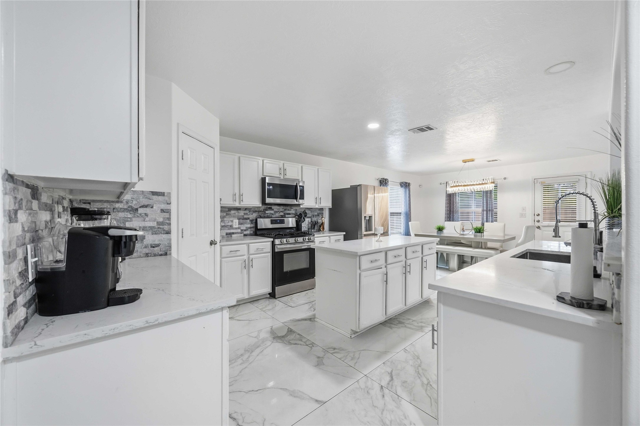 3538 Maris Way Humble, TX 77338 - Photo 8 of 24 Stylish and spacious kitchen showcasing crisp white cabinetry, marble-look tile flooring, and a contemporary tile backsplash. The layout features a large center island for added prep space and storage, along with stainless steel appliances and ample counter space. Bright recessed lighting and an open flow to the living and dining areas make this kitchen both functional and ideal for entertaining.