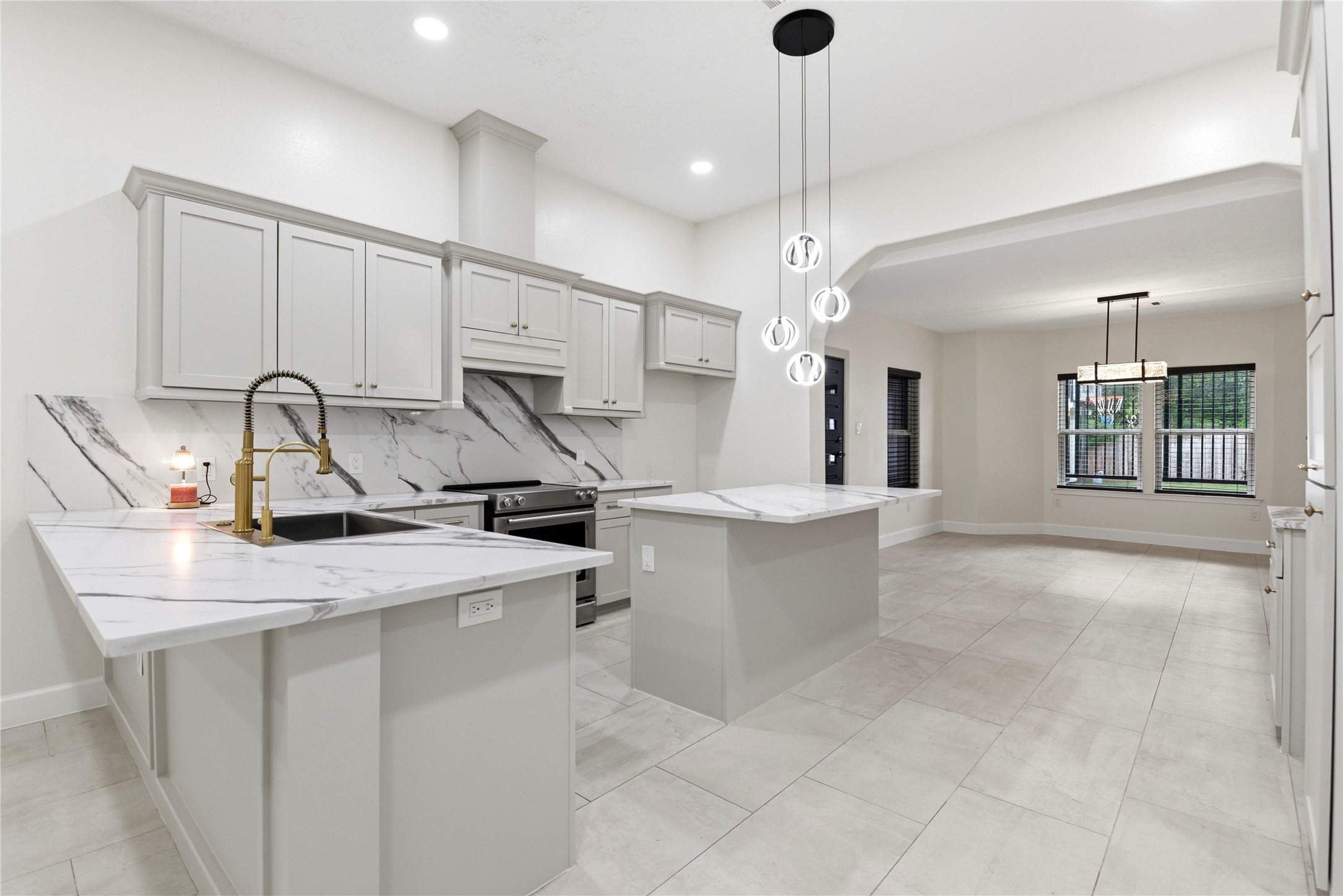 9403 Spaulding Street Houston, TX 77016 - Photo 11 of 50 This modern kitchen features sleek white cabinetry, marble countertops, and a matching backsplash. It includes stainless steel appliances, a central island with elegant pendant lighting, and an open layout leading to a bright dining area with large windows.